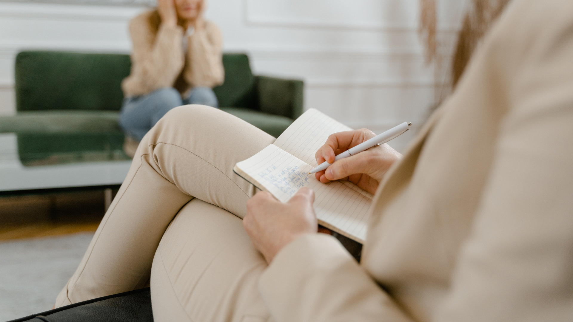 Therapist taking notes while a person on a couch covers their face in a neutral-toned room.