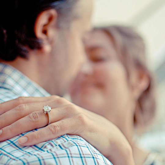 Man embraces woman, engagement ring on her finger, focus on ring.