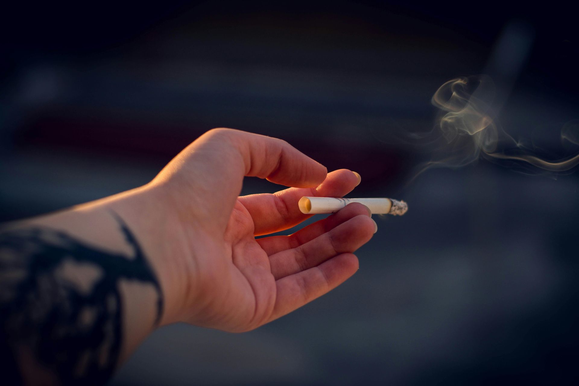 Hand with tattoo holding a lit cigarette, smoke rising, against a blurred background.