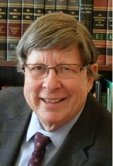 Man in suit and glasses smiling, bookshelves in the background.
