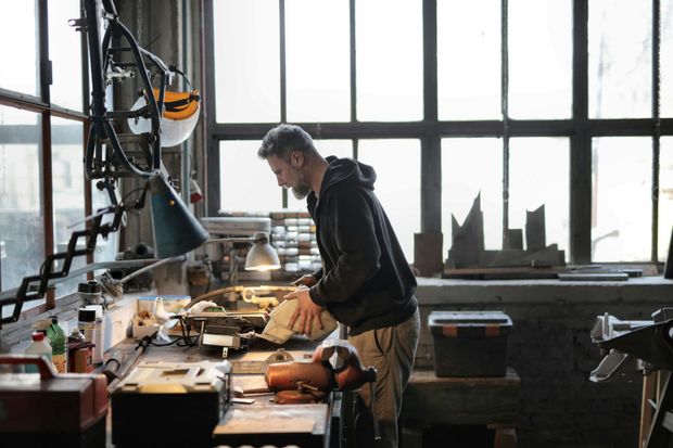 Man working at a workbench in a workshop. He wears a dark hoodie and beige pants, working in front of a large window.
