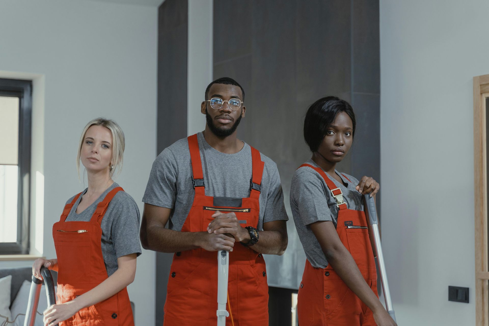 Three cleaning crew members, wearing red overalls and gray shirts, stand in a room holding cleaning supplies