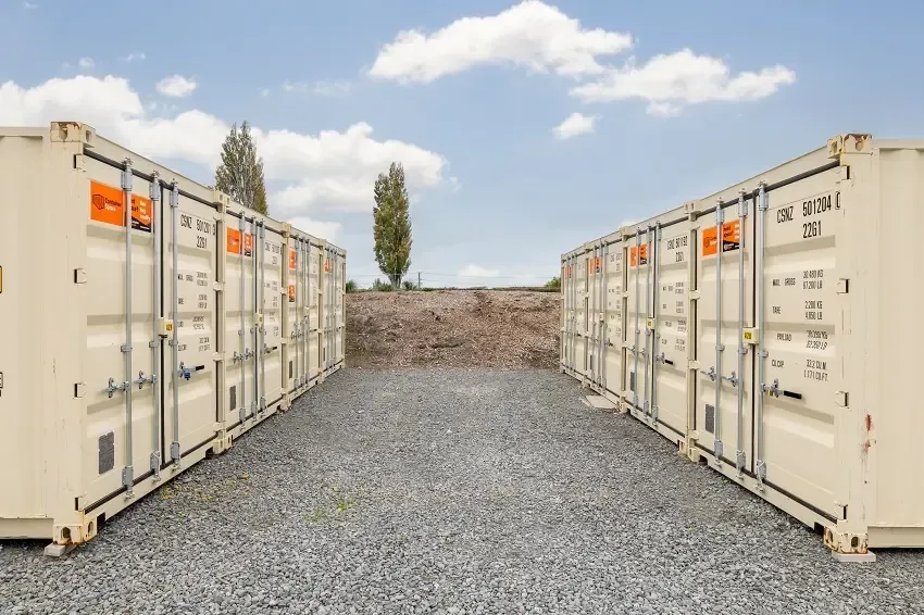 Two beige storage containers on a gravel lot, with a backdrop of trees and a blue sky.