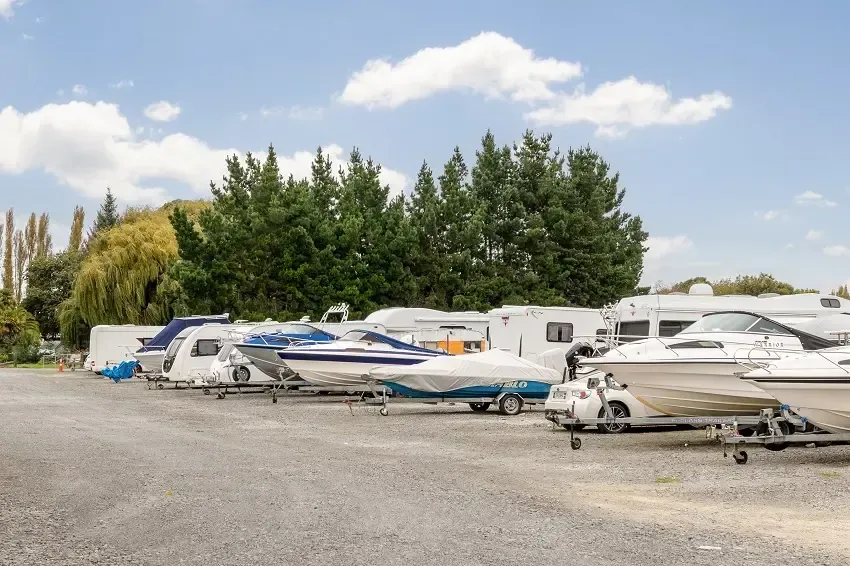 Boats and RVs parked in a gravel lot on a sunny day with trees in the background.