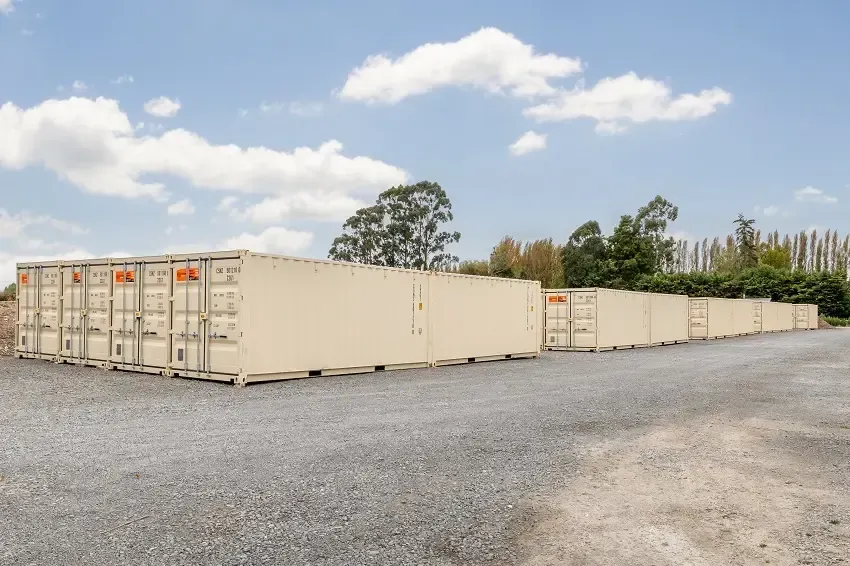 Rows of tan storage containers on a gravel lot under a partly cloudy sky. Trees in background.