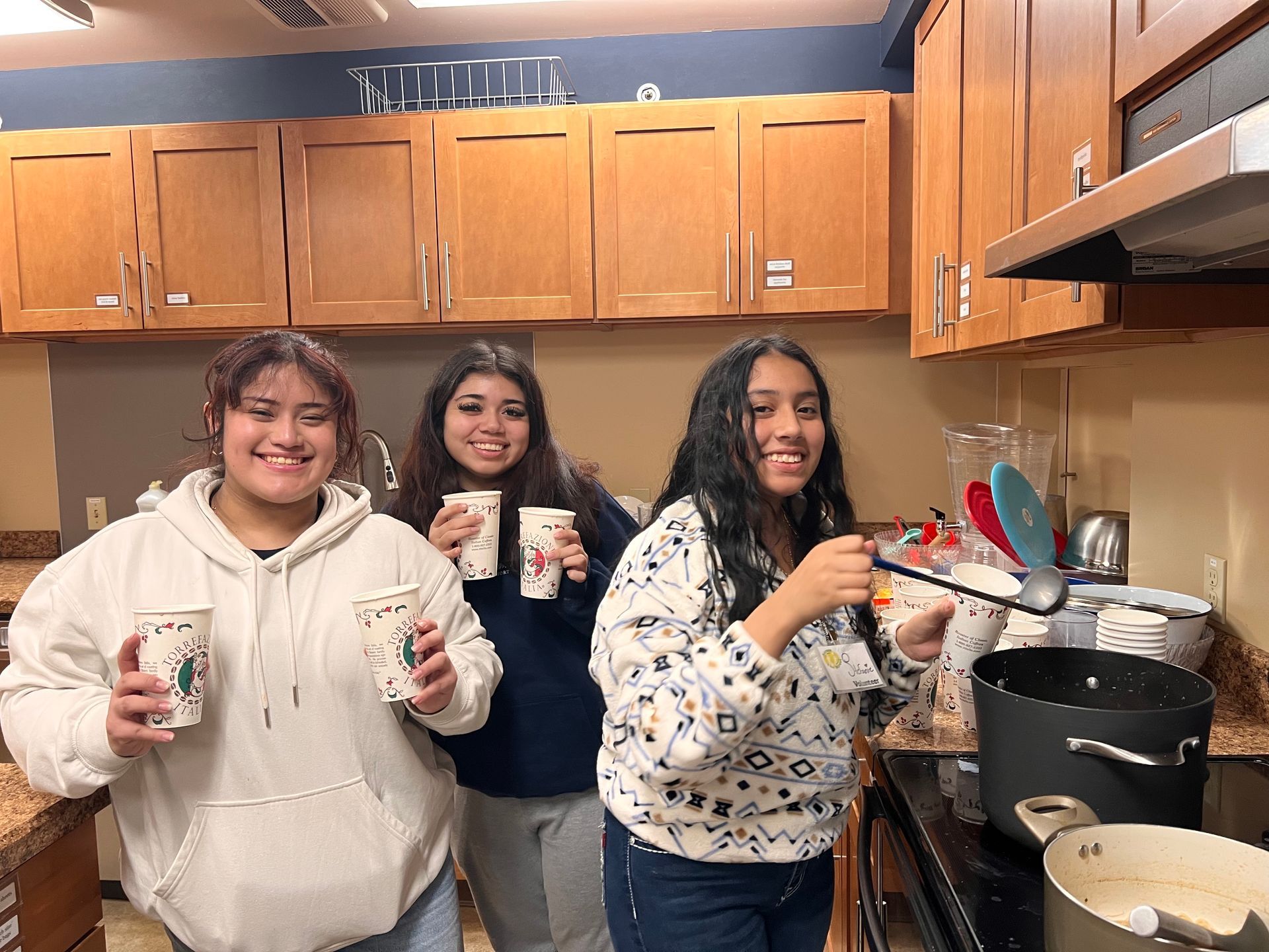 Three girls are standing in a kitchen holding cups and spoons.
