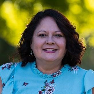 A  Latina woman in a blue floral shirt is smiling for the camera.