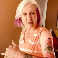 A blond woman with purple stripe in her hair and a tattoo on her arm is giving a thumbs up.