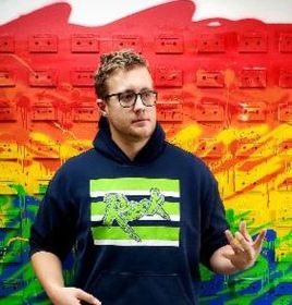 A man wearing glasses and a hoodie is standing in front of a rainbow wall.