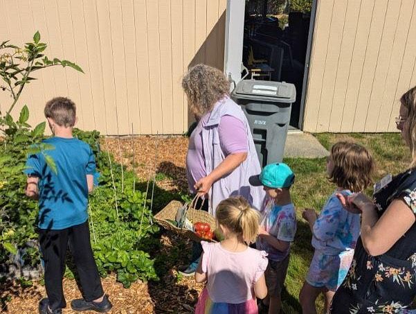 A group of children are standing in a garden with a woman holding a basket of tomatoes.