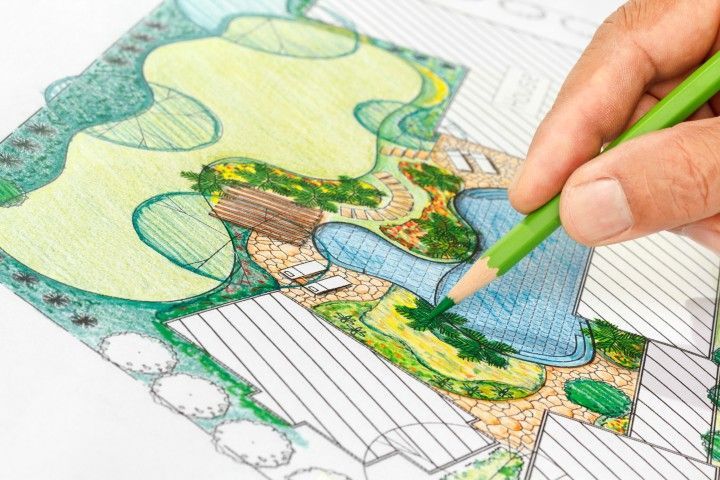 A hand uses a green pencil to color a detailed architectural landscape design plan featuring a pool and garden areas.