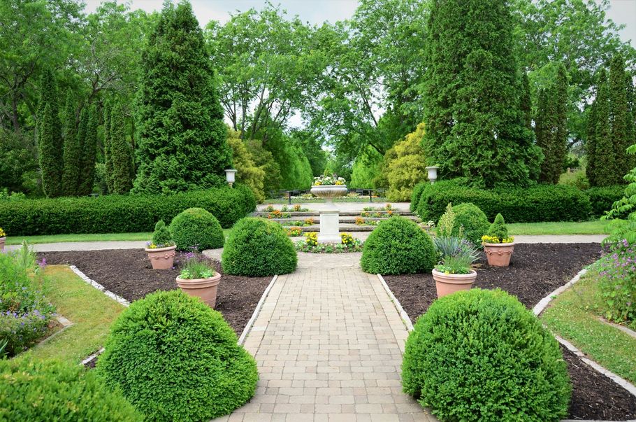 A stone path leads through a formal garden with manicured boxwood shrubs, potted plants, and tall, evergreen trees.