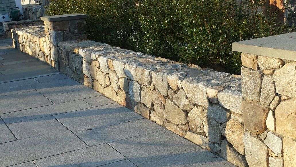 A stone wall along a sidewalk next to a bush.