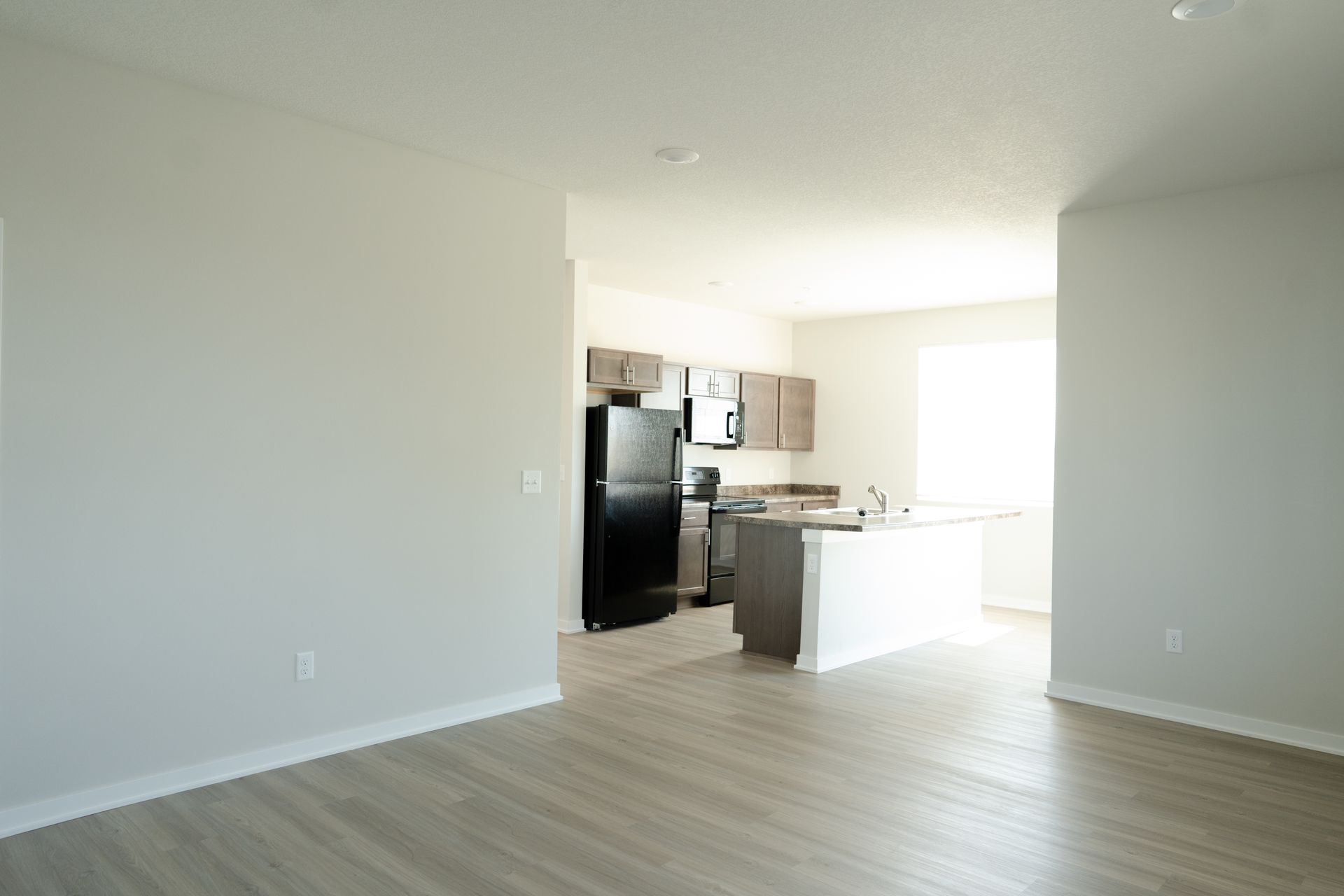 An empty living room with a black refrigerator and a kitchen in the background.