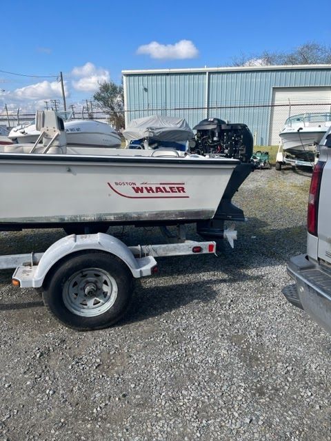 White Boston Whaler boat on a trailer, parked on gravel. A black motor is attached.