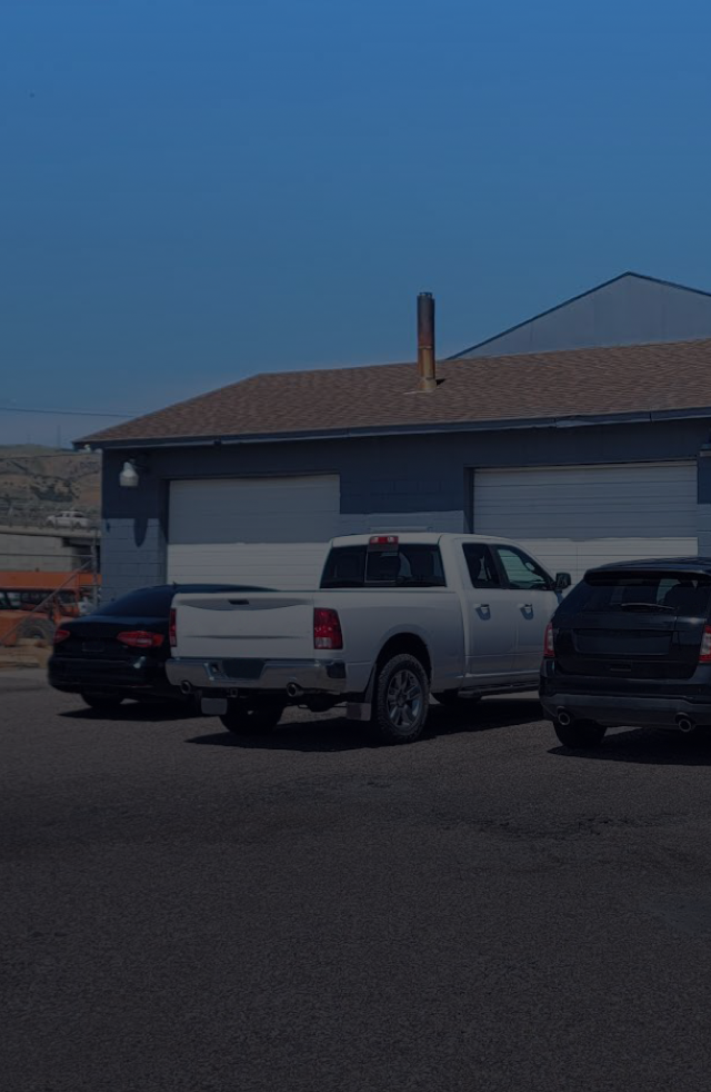 A white pickup truck parked in front of a gray building with garage doors, accompanied by a black car and SUV under a blue sky | Know A Guy Auto Repair