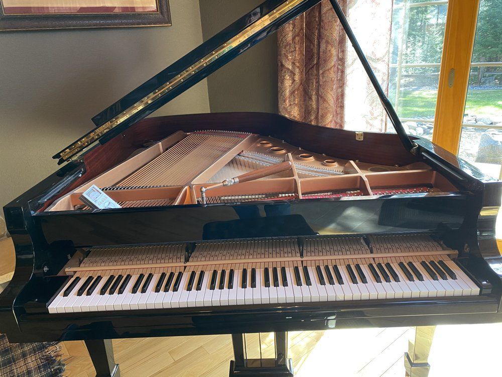 Black grand piano with its lid open, revealing strings and keys. Set near a window.