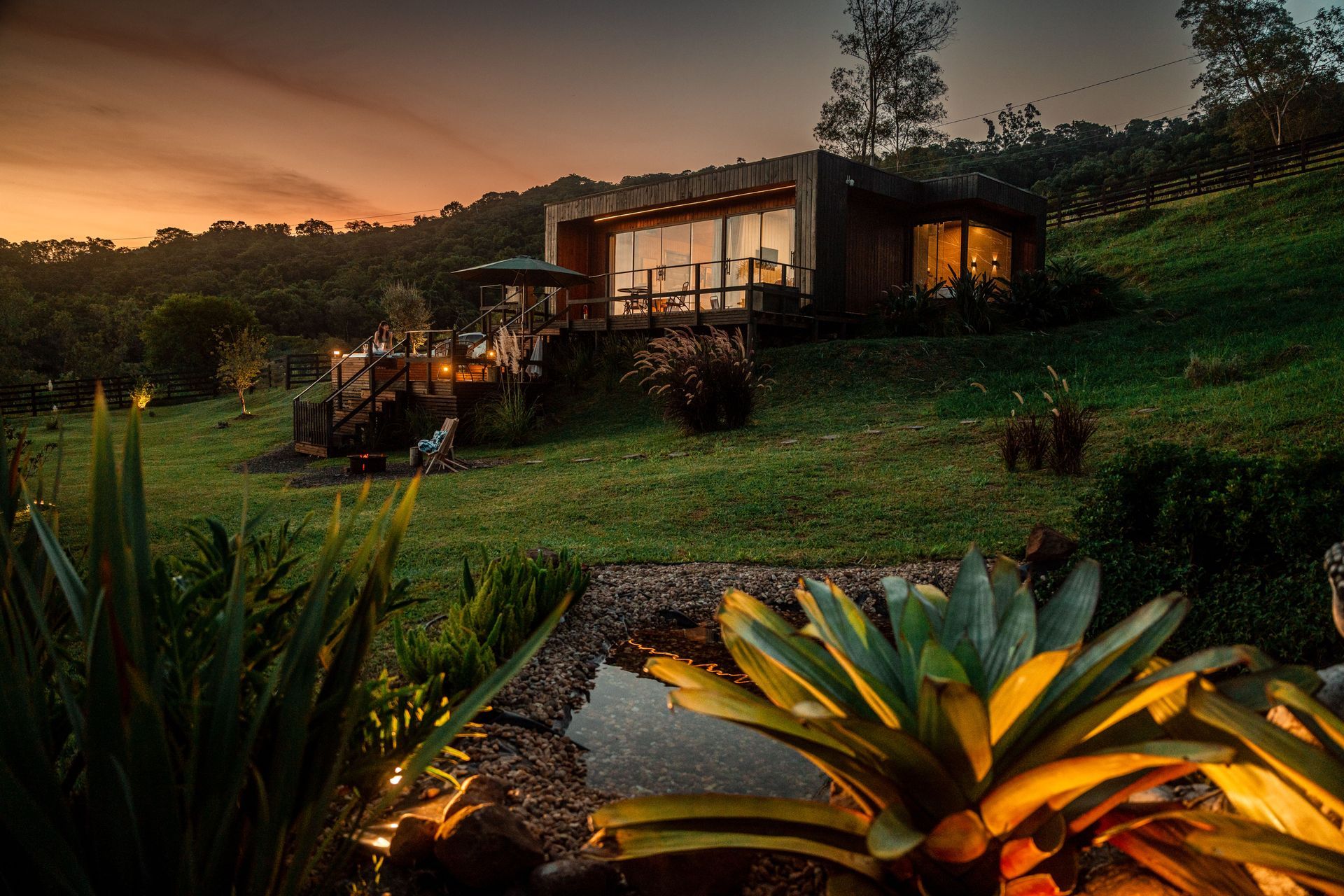 Uma cabana ao pôr do sol, rodeada de vegetação e um lago refletor.