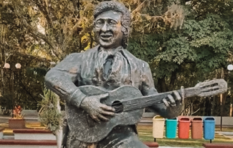 Estátua de um homem tocando violão em um parque. Árvores e latas de lixo coloridas estão ao fundo.