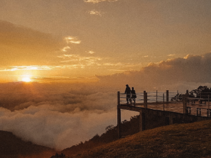 Vista do pôr do sol a partir de um mirante acima das nuvens; silhueta de duas figuras, luz dourada.