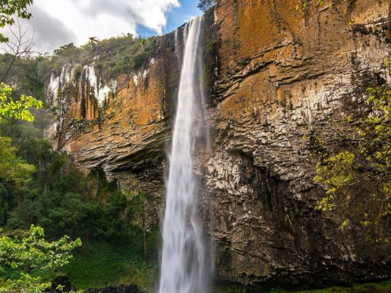 Cachoeira que despenca por uma encosta rochosa acidentada e bege, com folhagem verde
