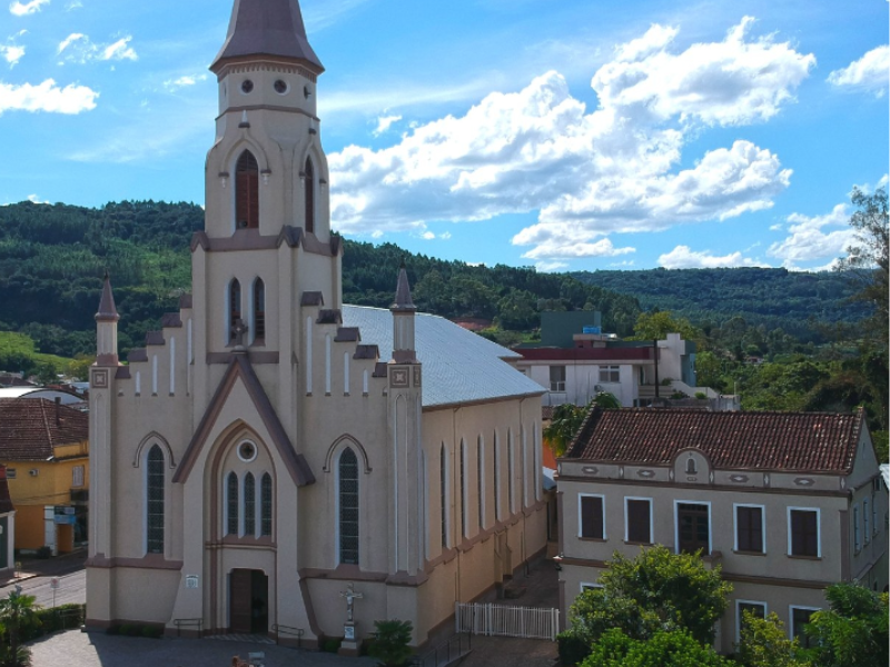 Igreja com torre alta, fachada bege clara, tendo como pano de fundo árvores verdes e céu azul.