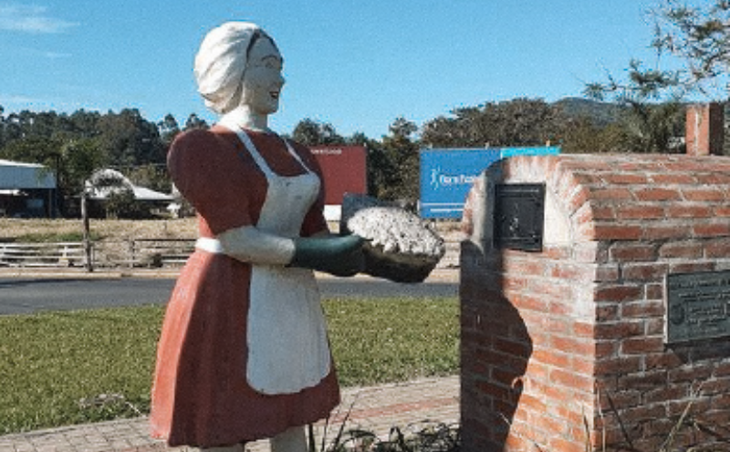 Estátua de mulher de avental segurando uma torta, em pé ao lado de uma estrutura de tijolos com uma placa, 