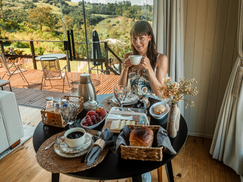 Mulher desfrutando do café da manhã com uma bela vista; mesa posta com doces, frutas e café.