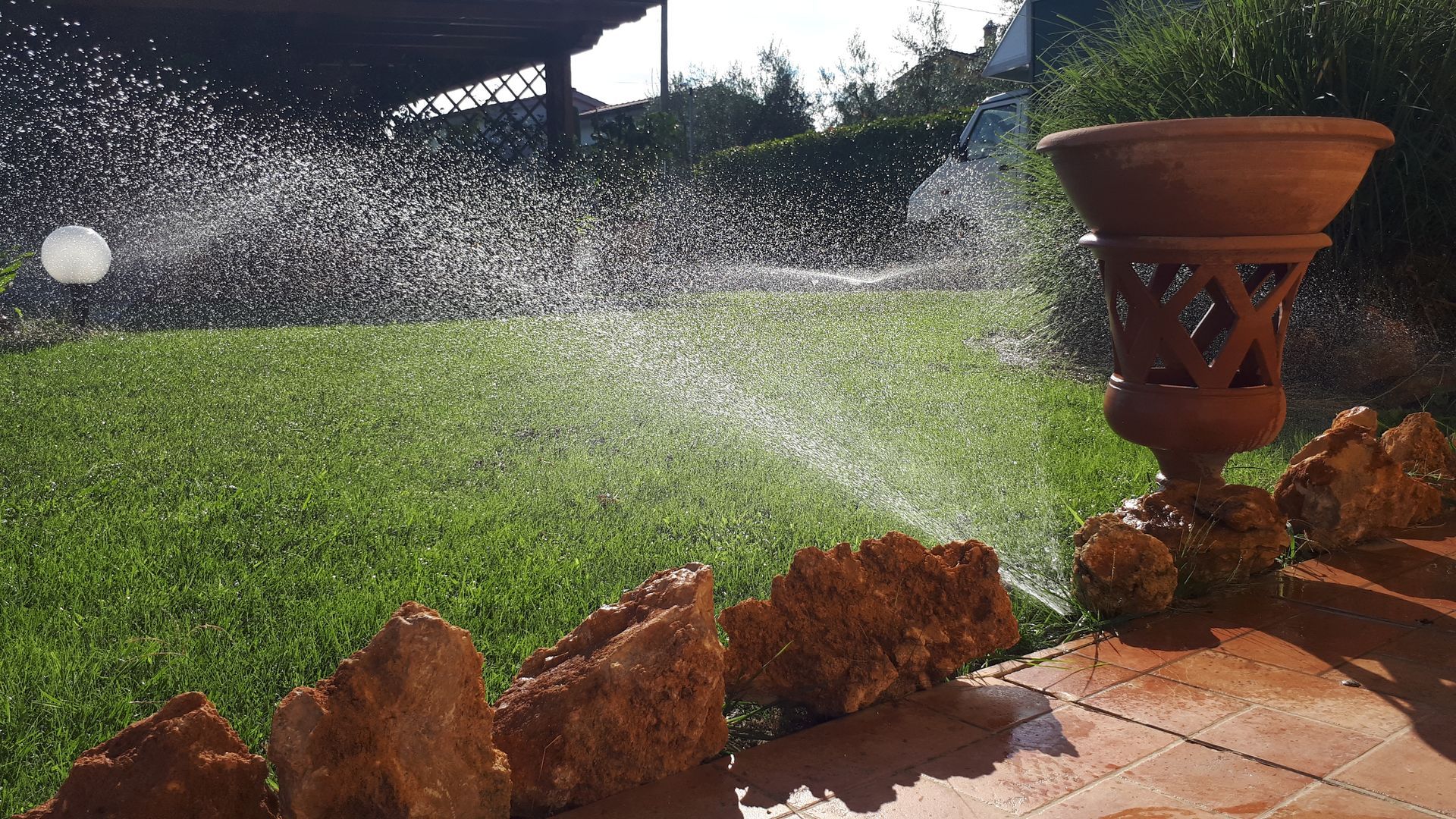 Irrigatore da giardino che spruzza acqua sull'erba verde in un cortile con una bordura decorativa in pietra e un vaso di terracotta.