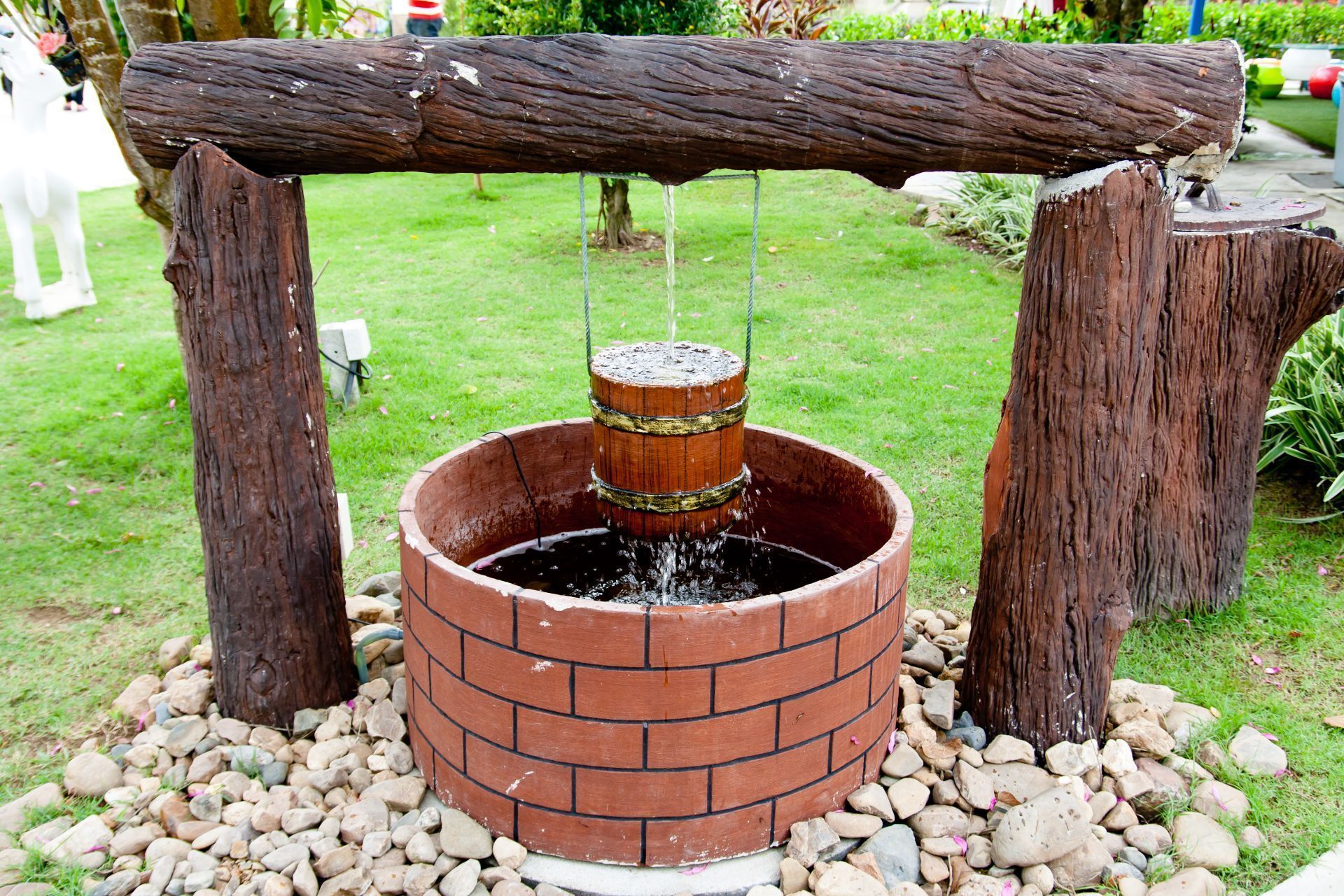 Pozzo decorativo con apertura rivestita in mattoni, secchio di legno e supporti in tronchi, situato in un cortile erboso.
