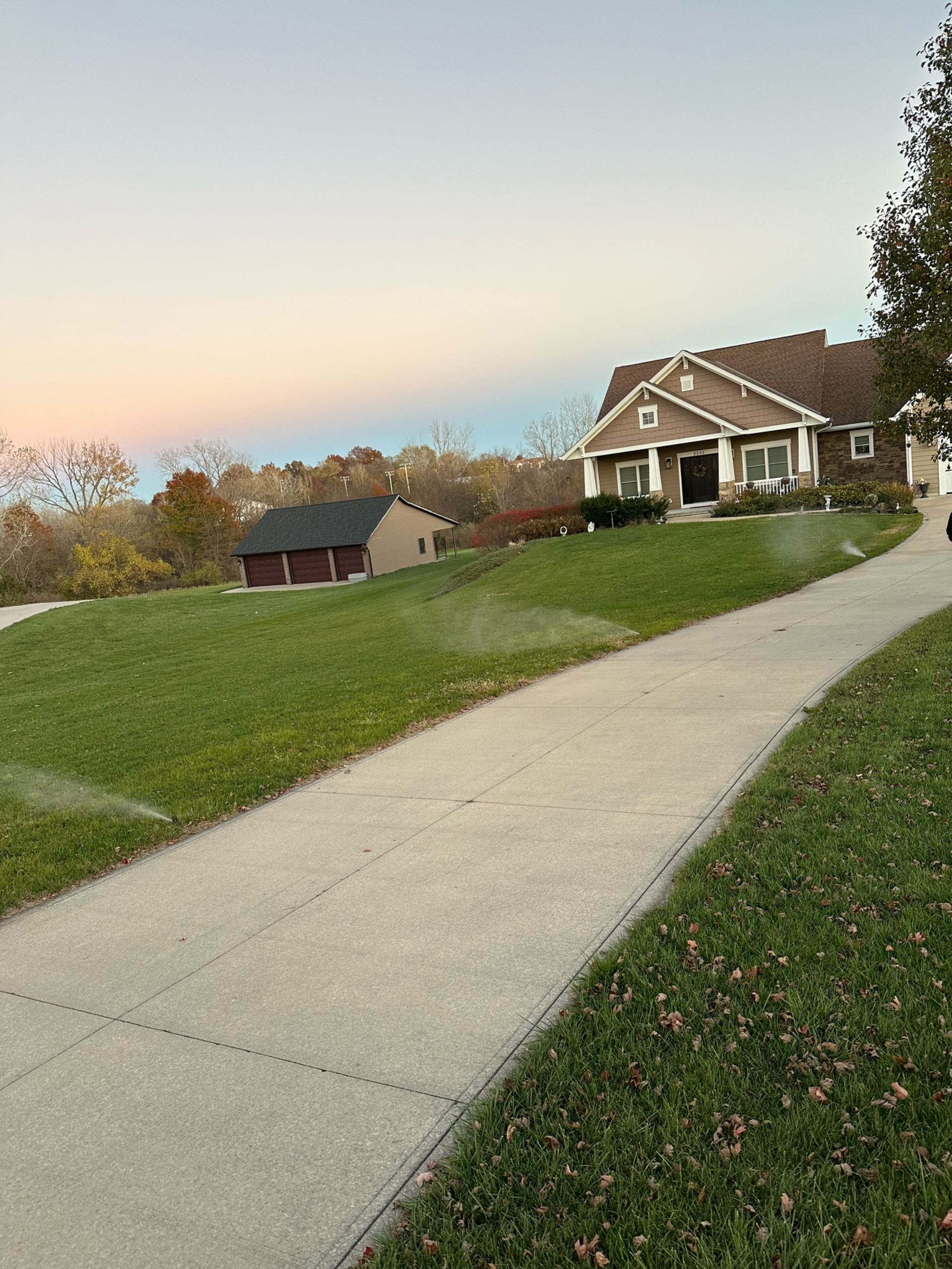 Sidewalk leading to a house on a hill with green grass and a shed. Sky is light blue and pink.