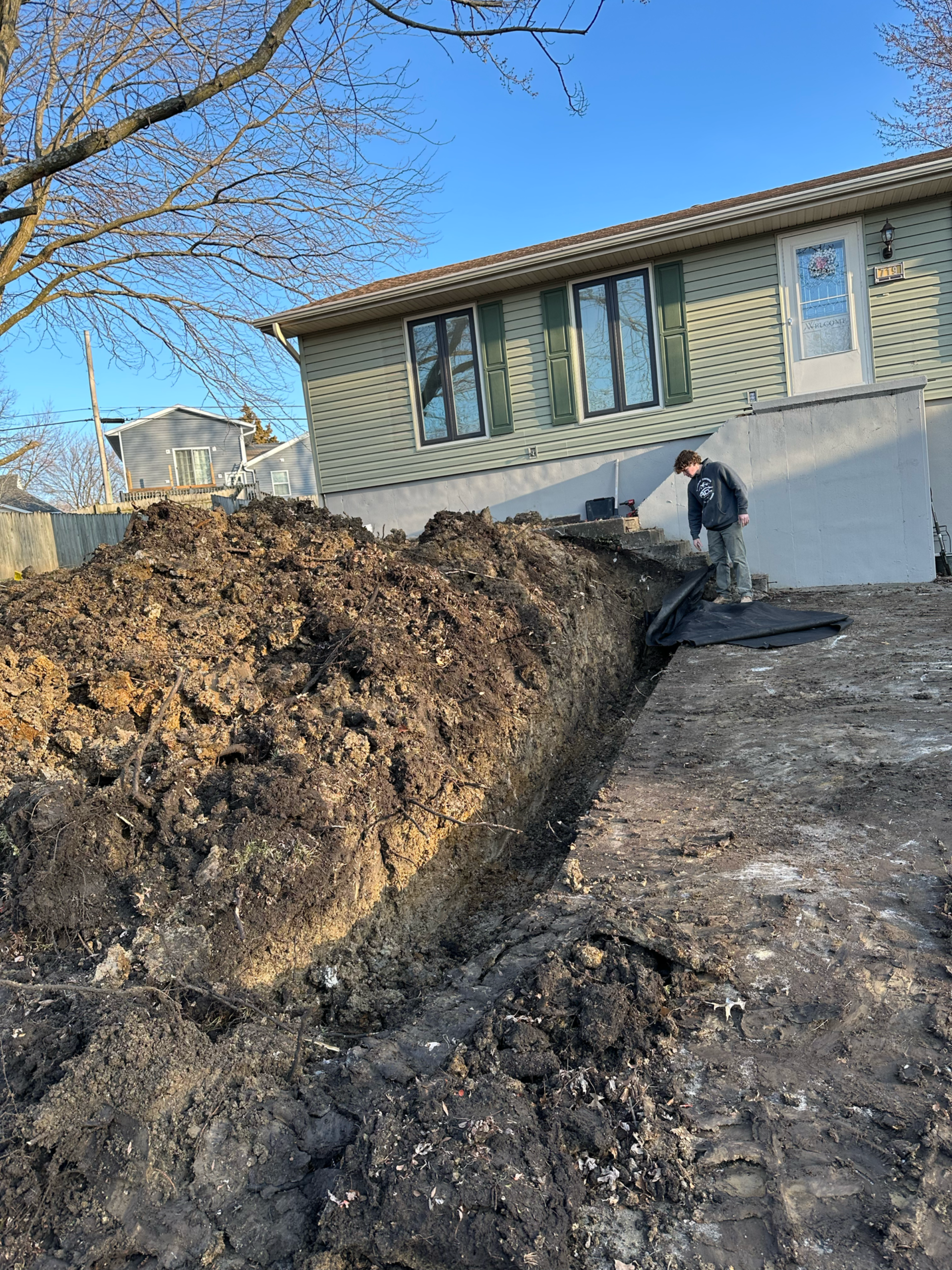 A man stands near an excavated trench beside a house. Brown dirt piled up.