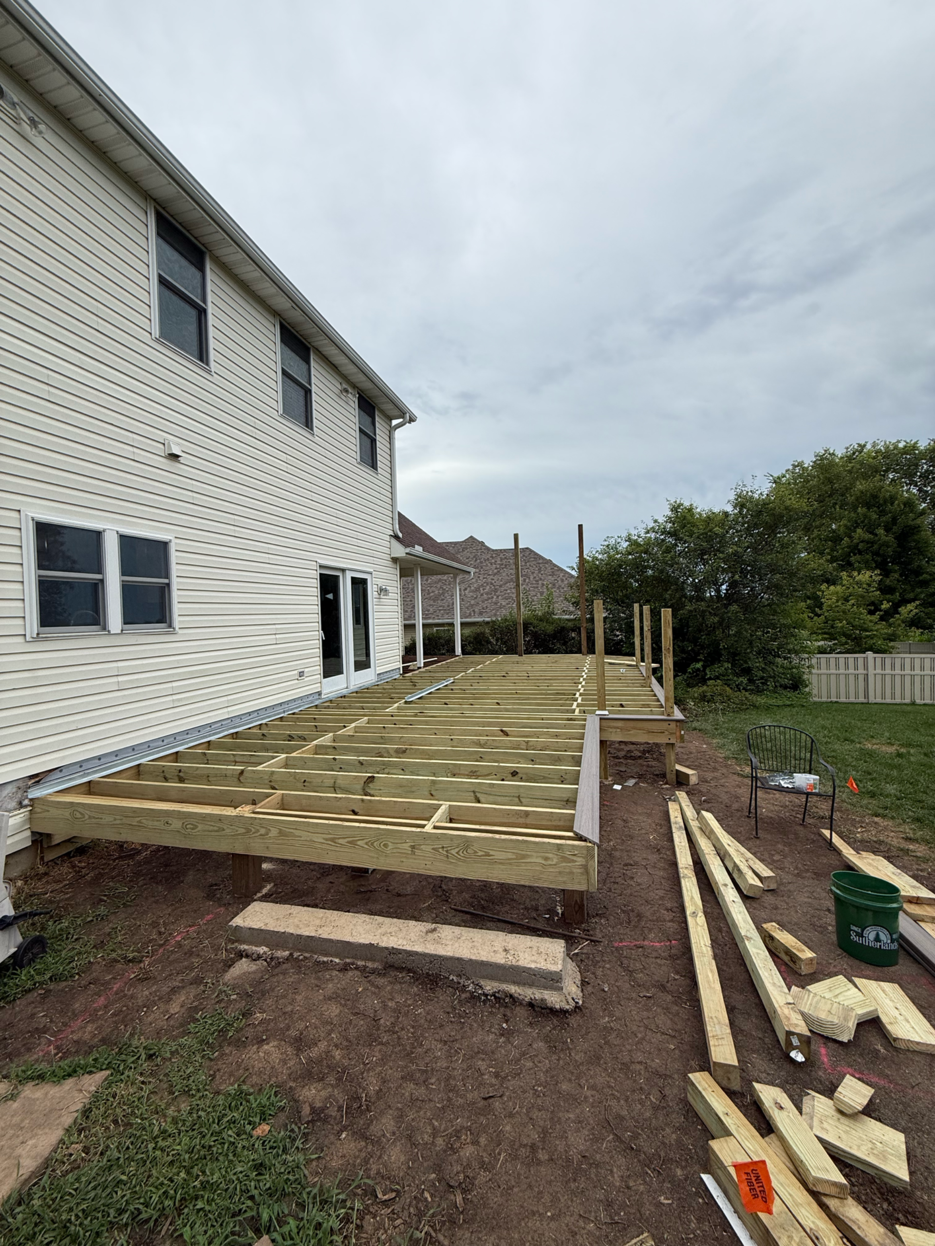 Deck construction in progress next to a two-story house. Unfinished wooden frame with exposed support beams.