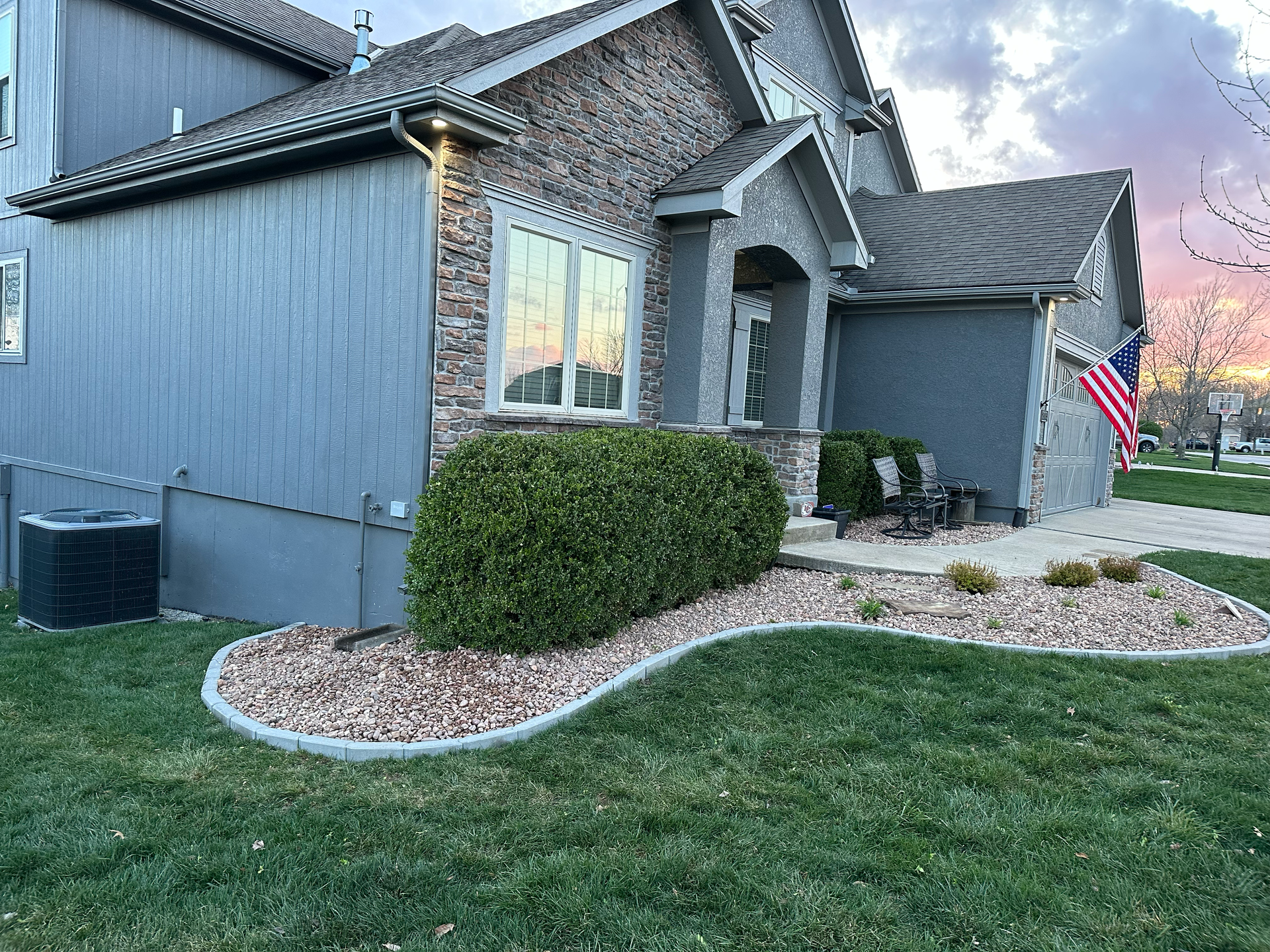 Gray house with stone accents, green lawn, and trimmed bushes; American flag waves.