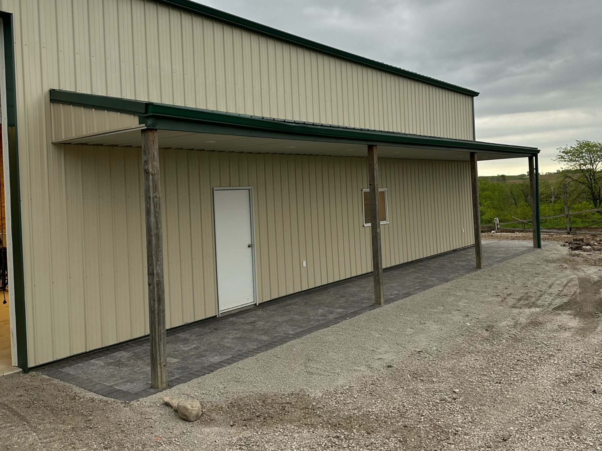 Tan metal building with a green awning supported by wooden posts; gravel ground.