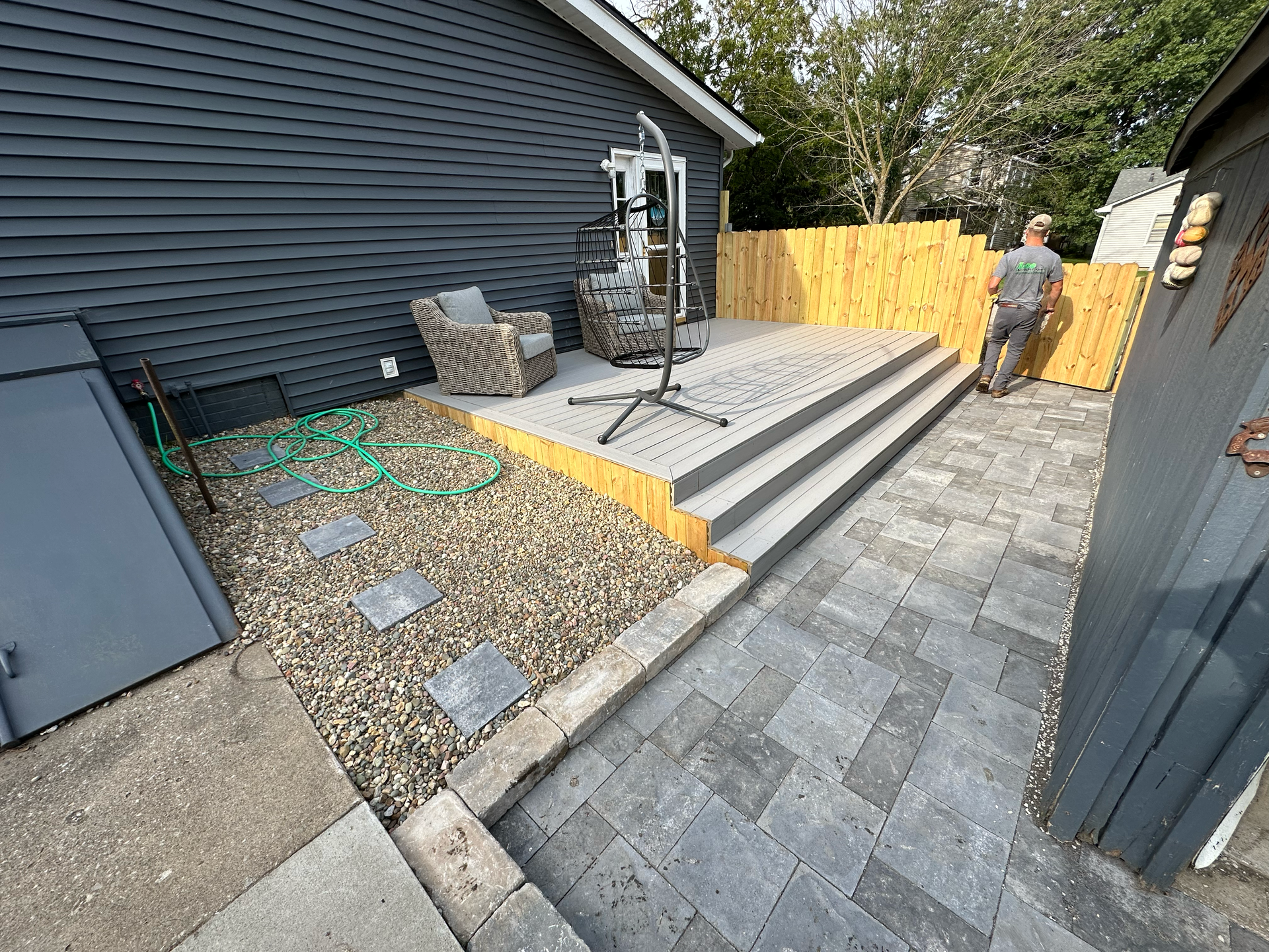 Backyard with grey composite deck, patio, gravel, and a person near a wooden fence.