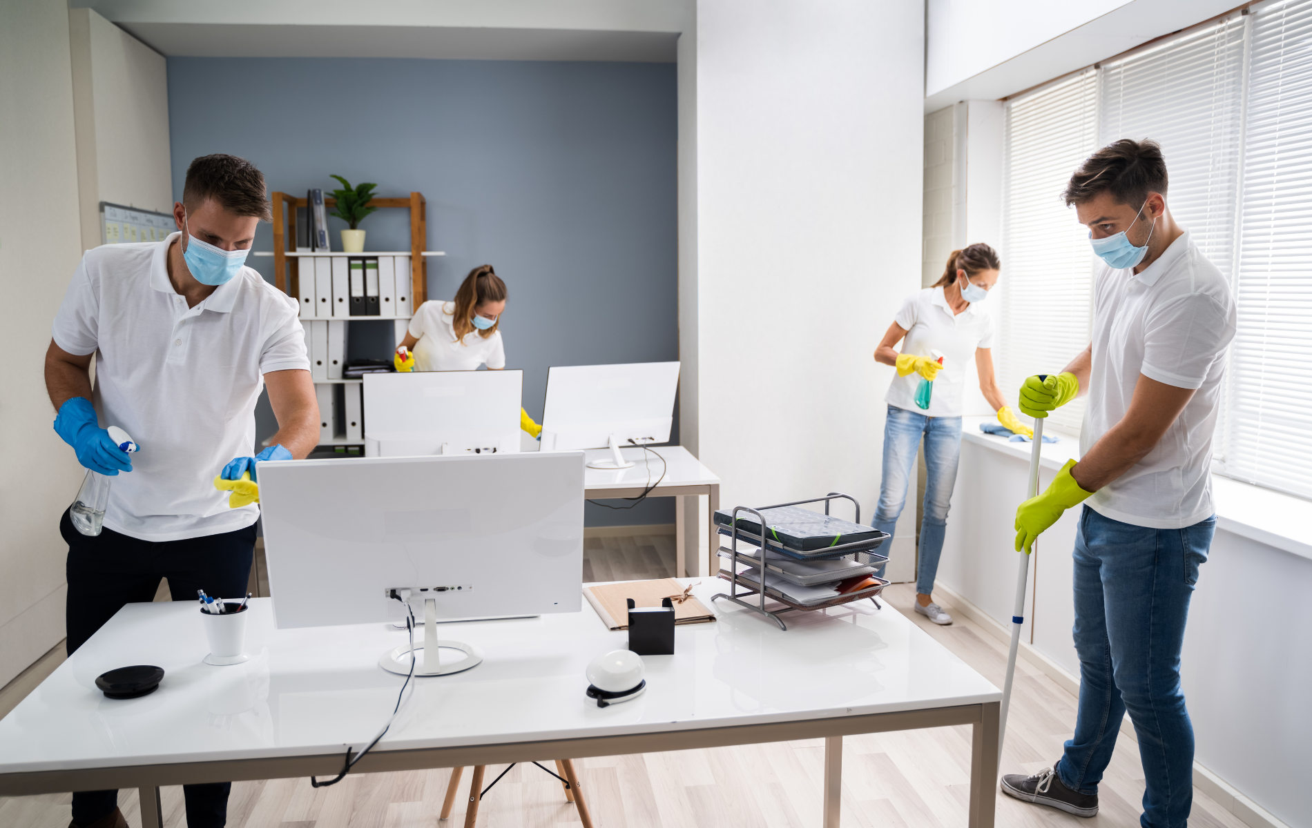 Four people wearing masks and gloves cleaning an office. Desks and windows are visible.