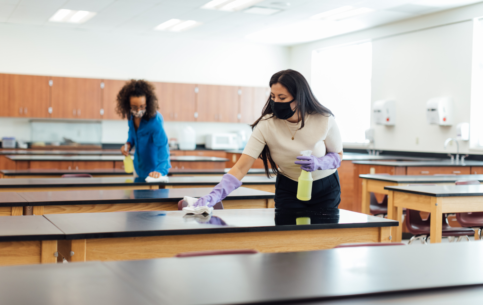 Two people wearing masks and gloves cleaning desks in a classroom.