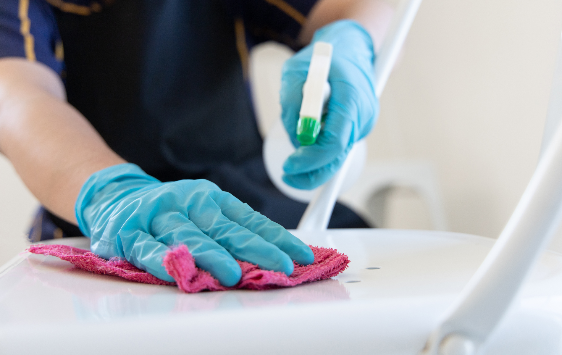 Person cleaning a white chair with a pink cloth and spray bottle, wearing blue gloves.