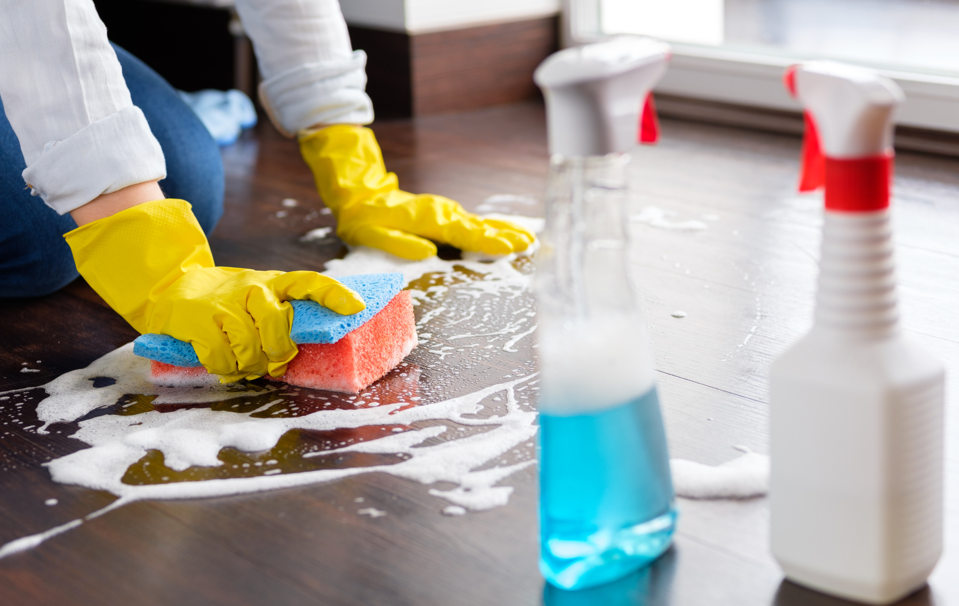 Person in yellow gloves cleaning wooden floor with a sponge and spray bottles.