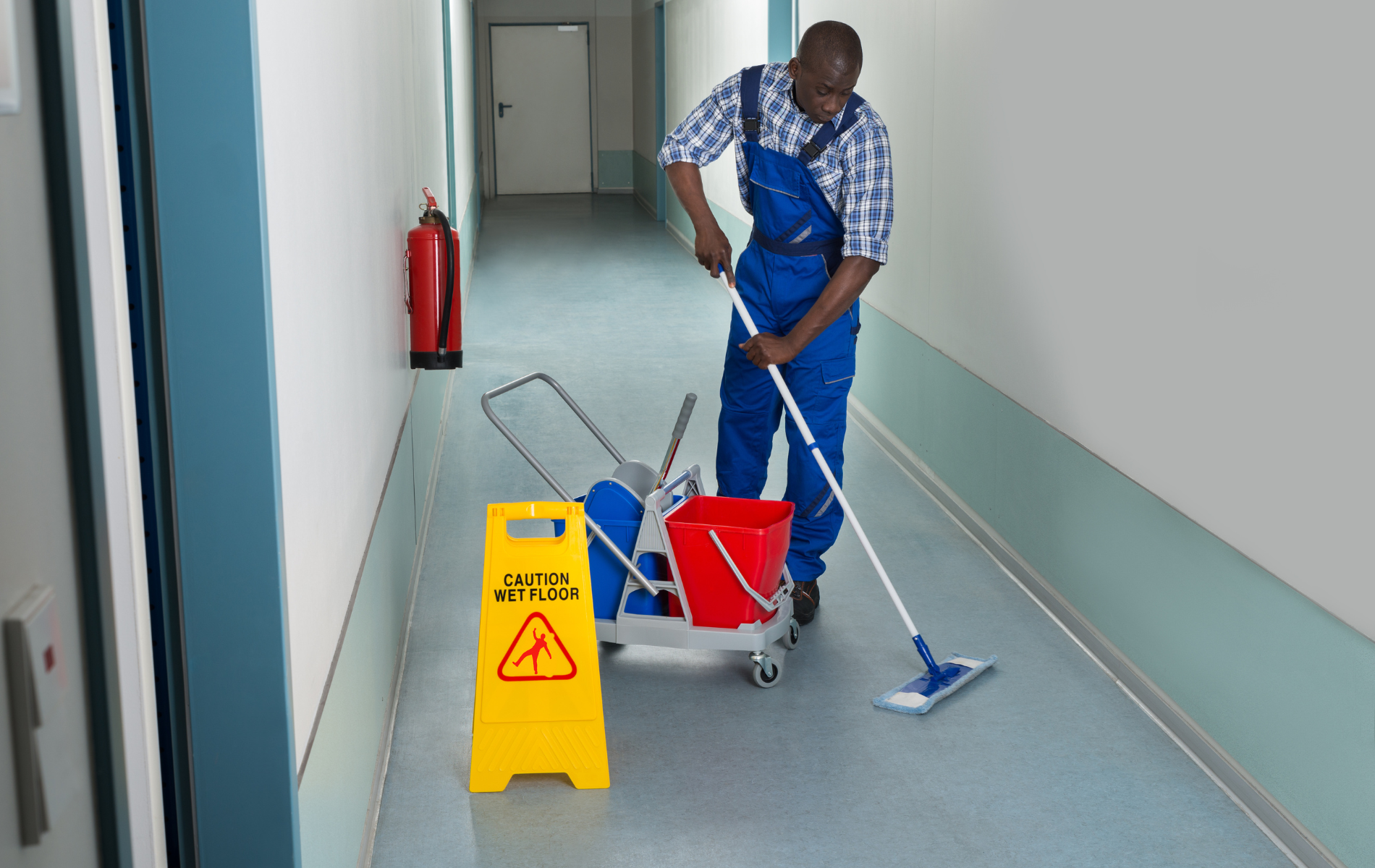Person mopping a hallway with a caution sign and cleaning supplies on a cart.