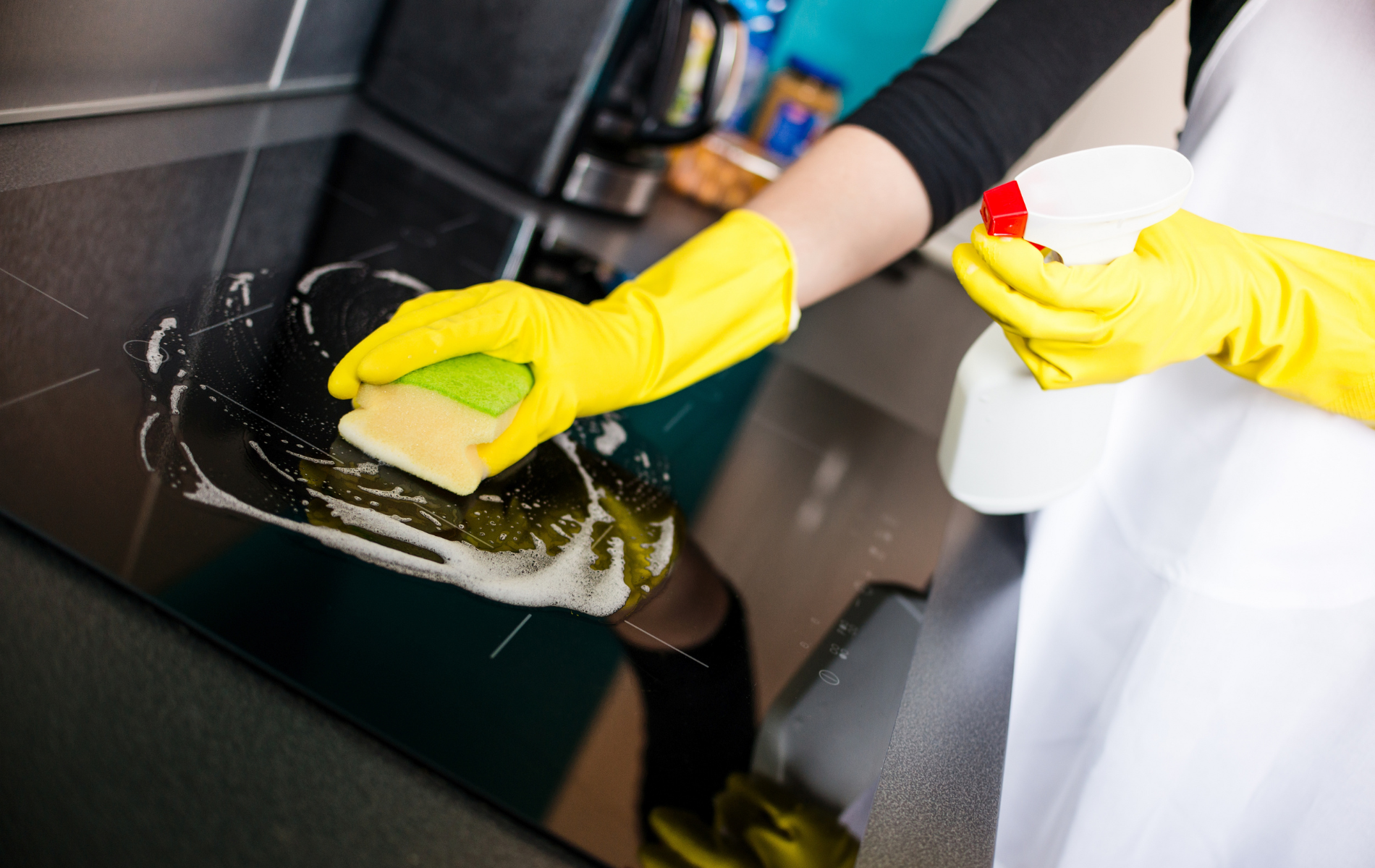 Person in yellow gloves cleaning a black stovetop with a sponge and spray bottle.