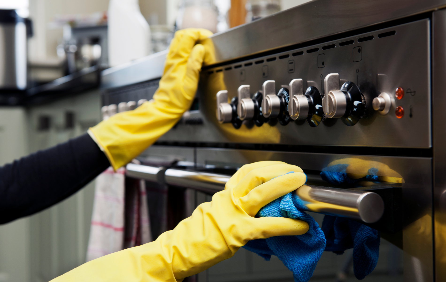 Hands in yellow gloves cleaning a stainless steel oven with a blue cloth.