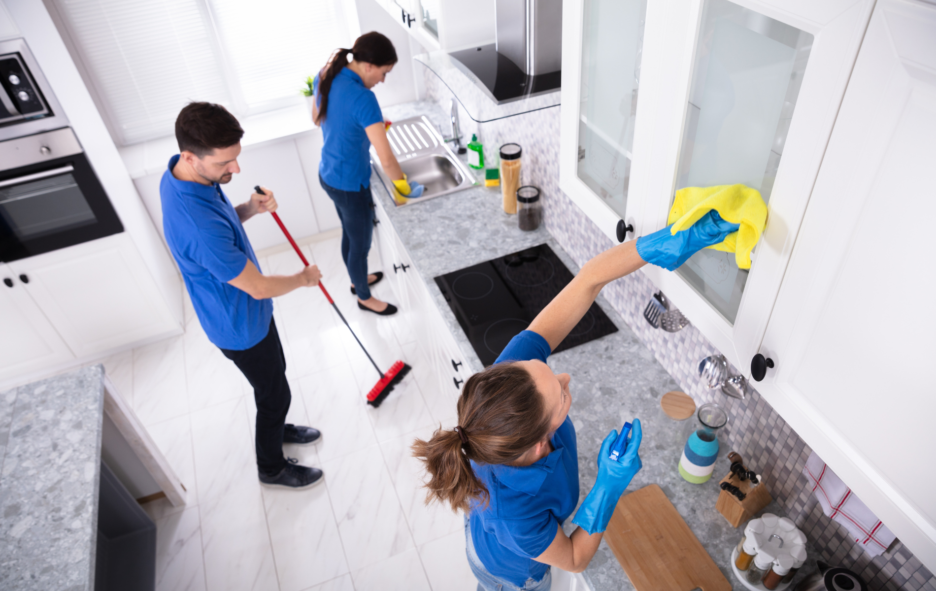 Three people cleaning a kitchen; one sweeps, one washes dishes, one wipes a cabinet.