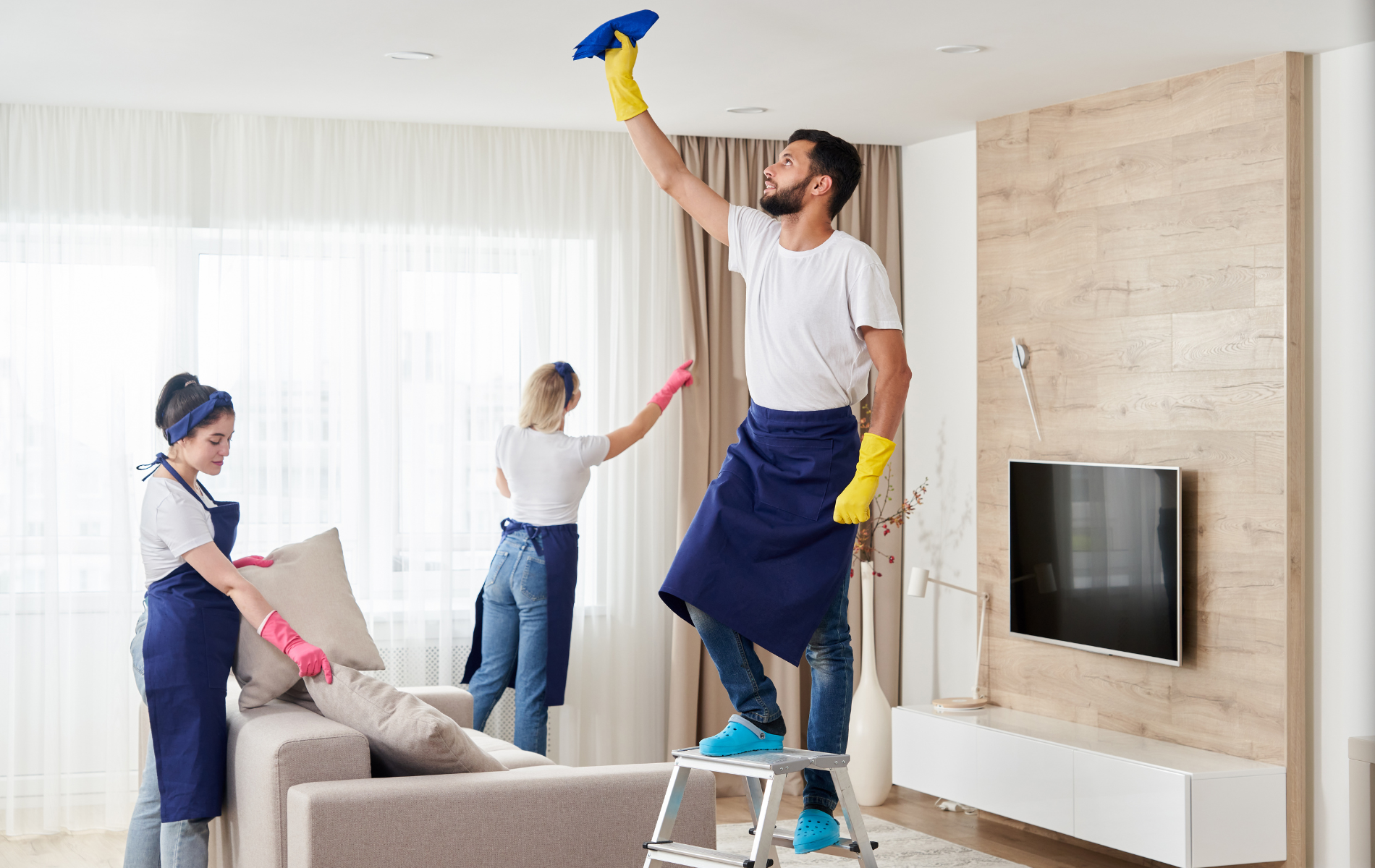 Three people cleaning a living room: one on a ladder cleaning the ceiling, two others tidying the sofa.