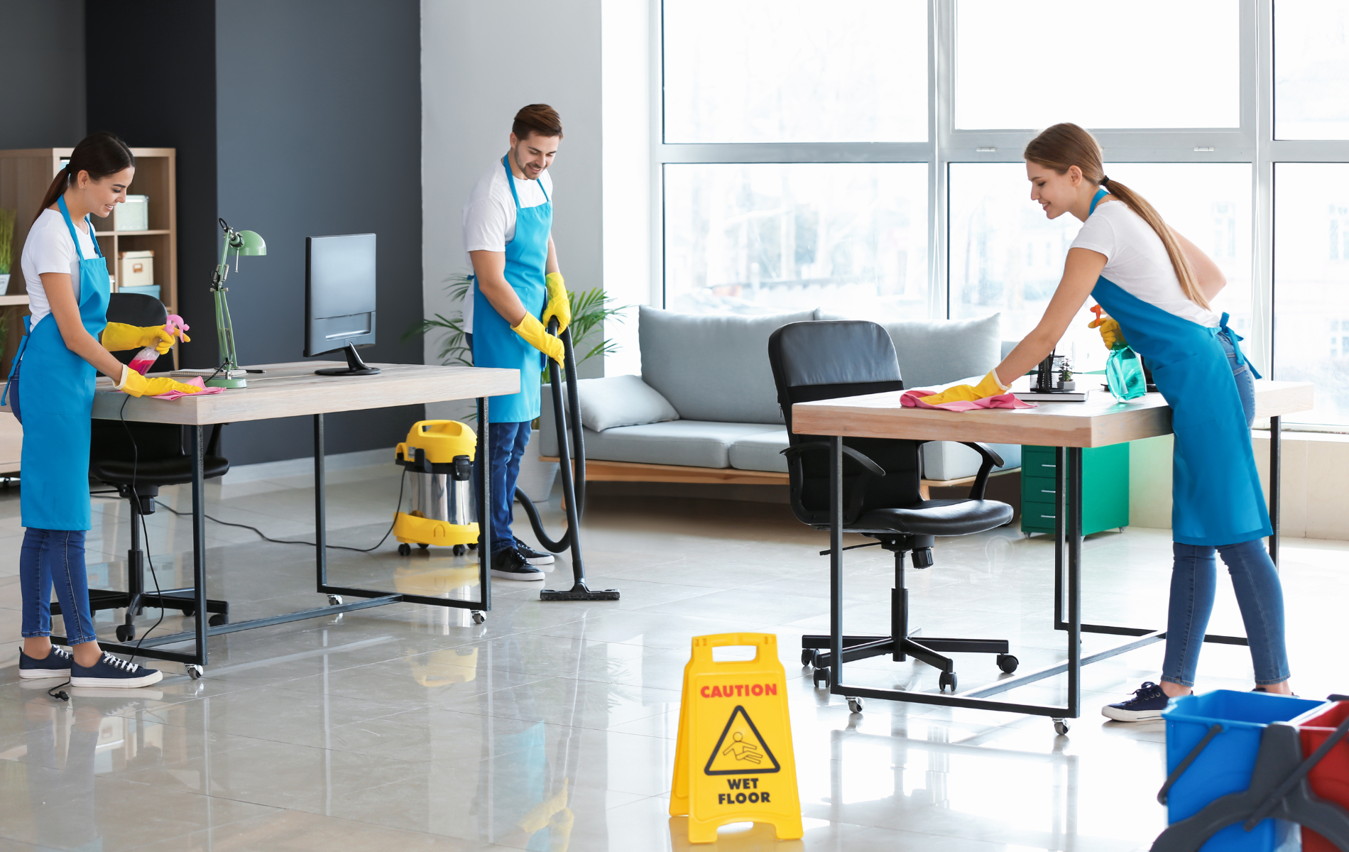 Three people cleaning an office: two wiping desks, one vacuuming. Yellow caution sign on the floor.