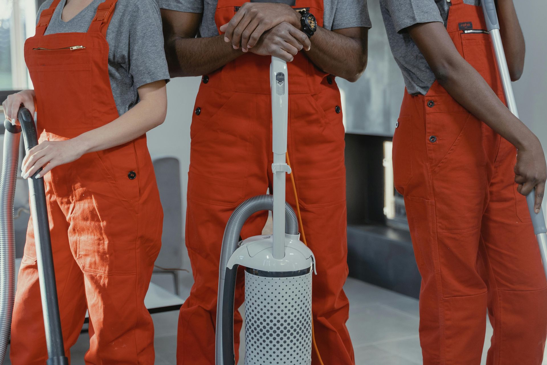Three people in orange overalls stand with a vacuum cleaner in a gray room.