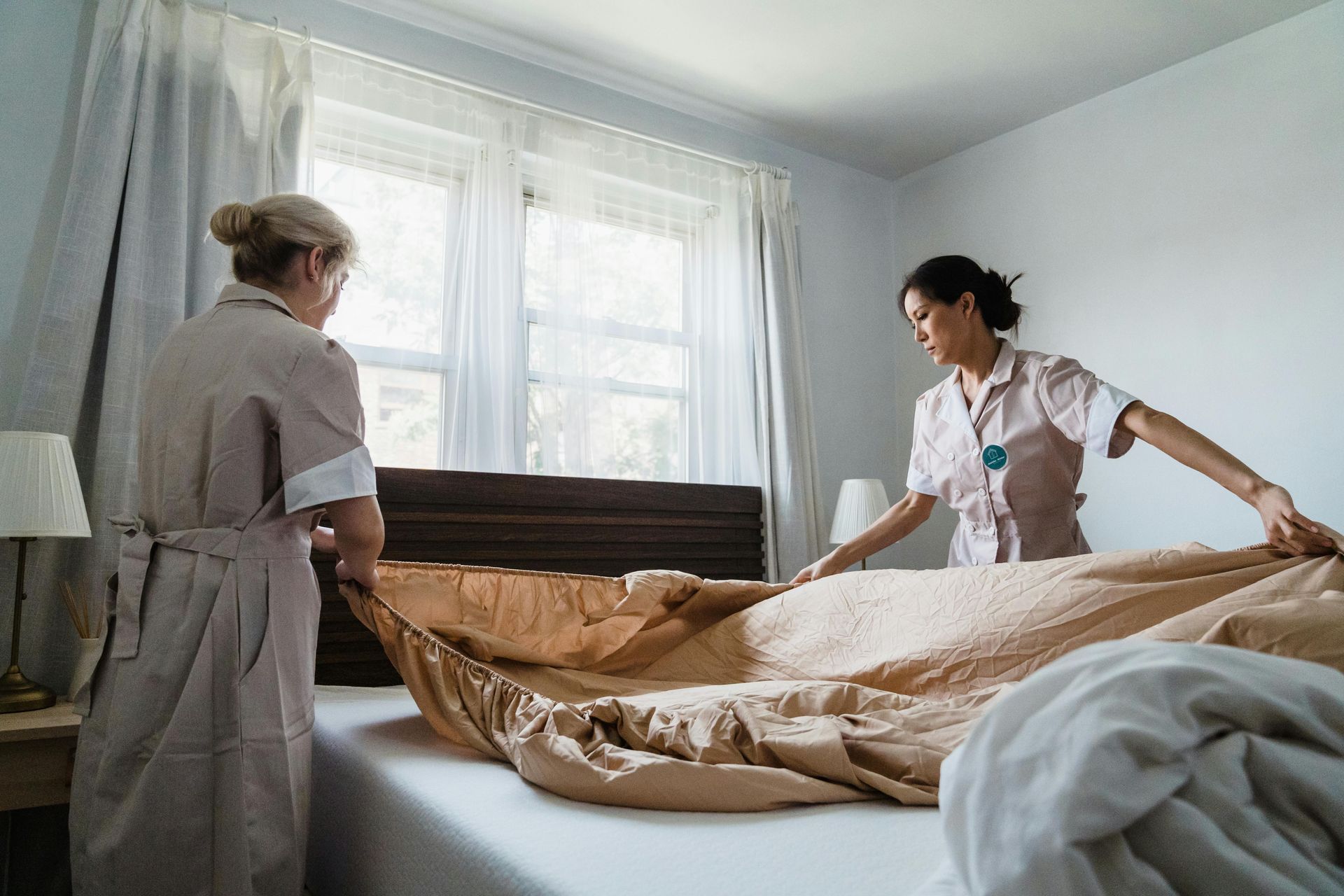 Two people in uniform making a bed in a room with a window and a nightstand.