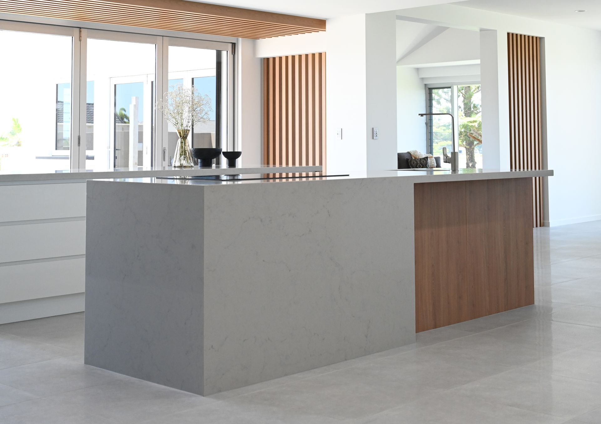 Modern kitchen with a gray countertop island and wood paneling. White cabinetry and flooring, with a sunny outdoor view.