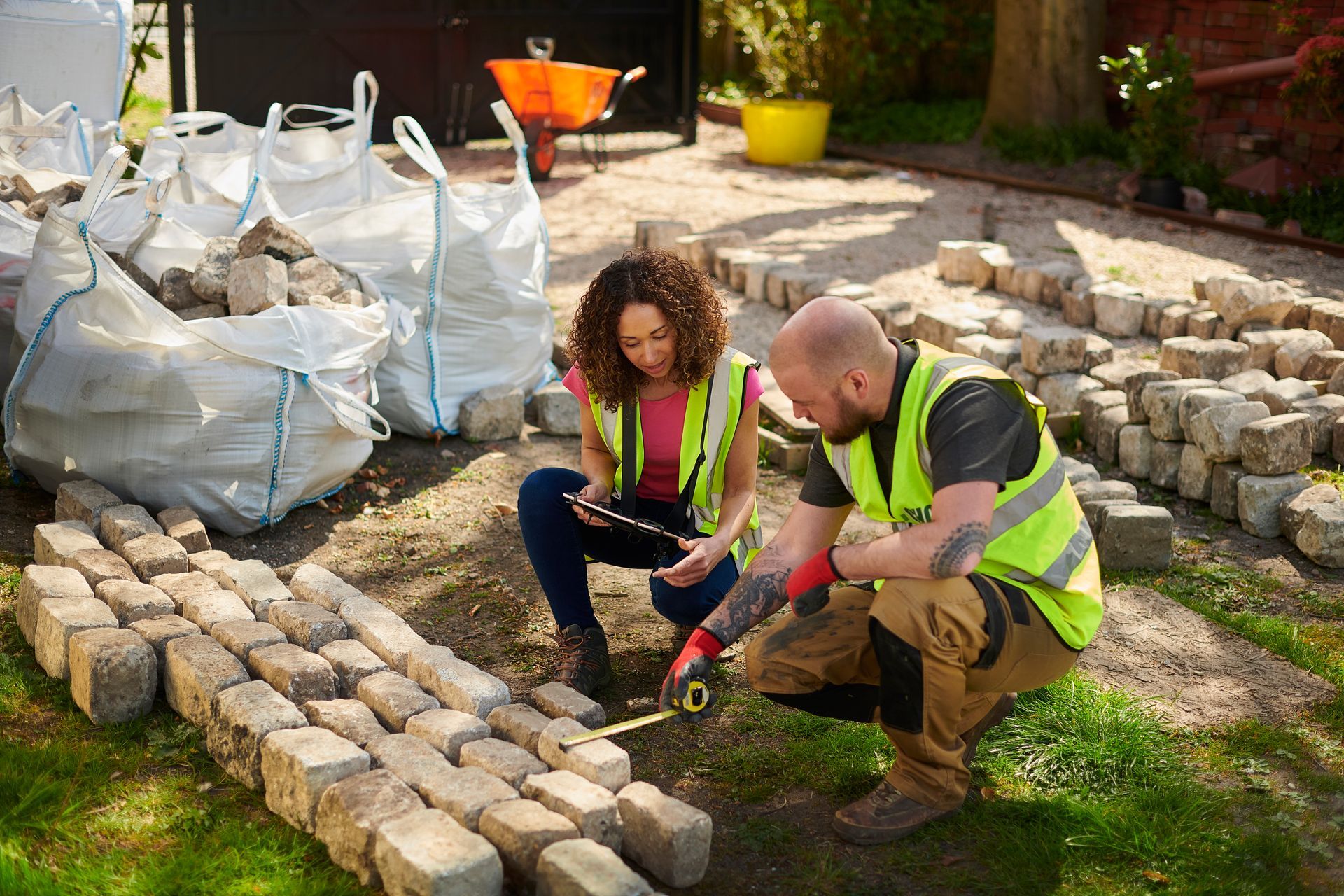 Two construction workers measure pavers outdoors, wearing vests.